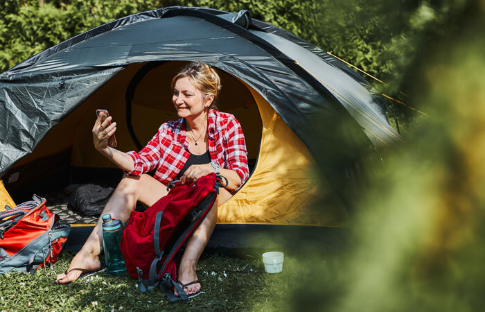 Female Having Video Call With Friends Using Smartphone While Sitting In Tent At Camping. Woman Relaxing In Tent During Summer Vacation. Actively Spending Vacations Outdoors Close To Nature. Concept Of Camp Life