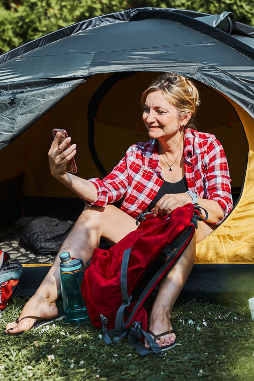 Female Having Video Call With Friends Using Smartphone While Sitting In Tent At Camping. Woman Relaxing In Tent During Summer Vacation. Actively Spending Vacations Outdoors Close To Nature. Concept Of Camp Life